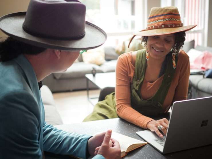 Shalom and Bo, two of RVC's capacity building leads, have a laptop and notebook open in between them as they converse. They are both dressed up slightly with fashionable accessories and hats.